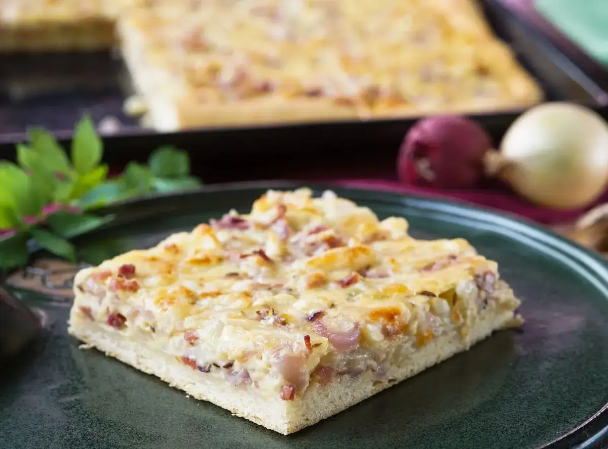 Ein saftiger Zwiebelkuchen vom Blech, mit Speck und Käse überbacken, liegt auf einem dunklen Teller.