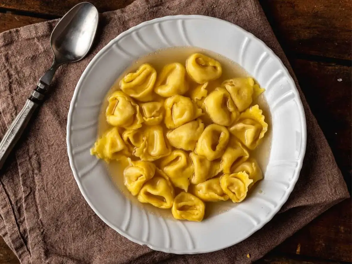 Un plato blanco lleno de deliciosos tortellini de carne en caldo, con una cuchara a un lado.