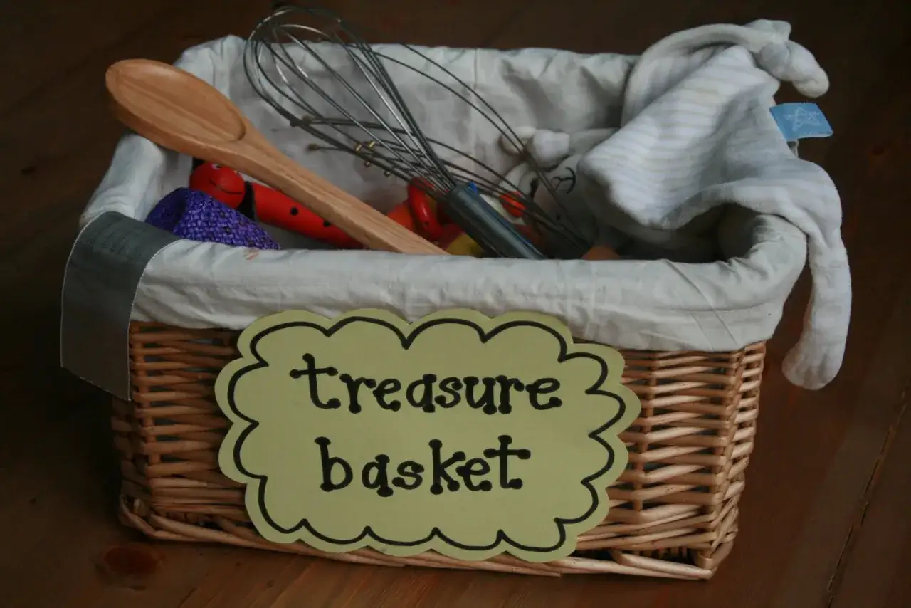 Cesta de mimbre con **juego heuristico materiales**: batidor, cuchara de madera, peluche y otros objetos.