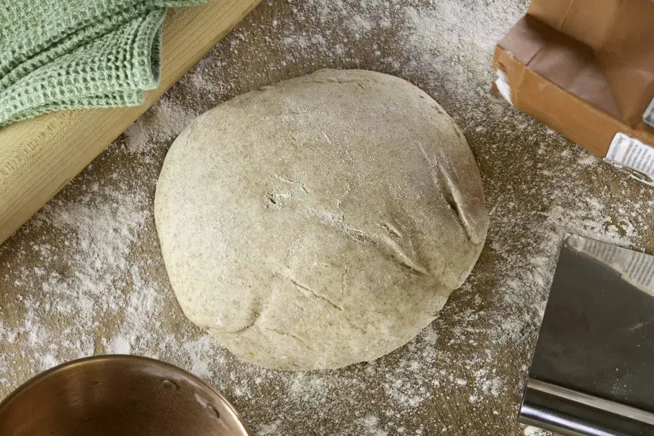 Pâte à pain sur une planche en bois saupoudrée de farine. Un rouleau à pâtisserie et un torchon vert sont visibles. La farine de riz complet bienfaits pour la santé sont nombreux.
