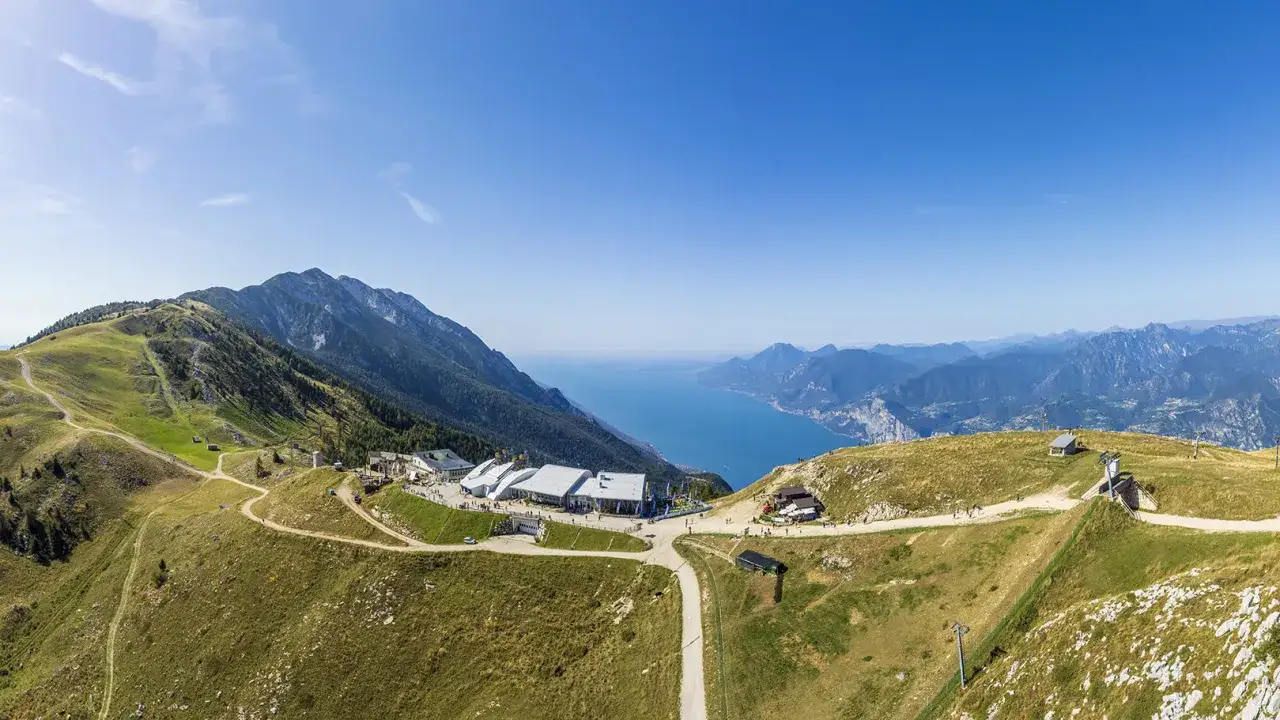 Panoramablick vom Monte Baldo auf den Gardasee. Geb&auml;ude und Wanderwege auf gr&uuml;nen H&auml;ngen unter blauem Himmel.