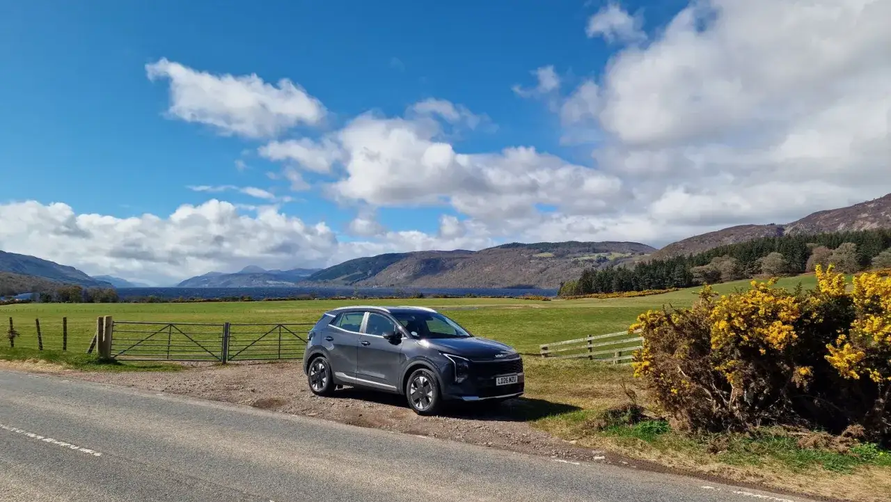 Coche gris aparcado junto a un lago escocés, ideal para viajar a Escocia por libre. Paisaje montañoso y cielo azul con nubes.