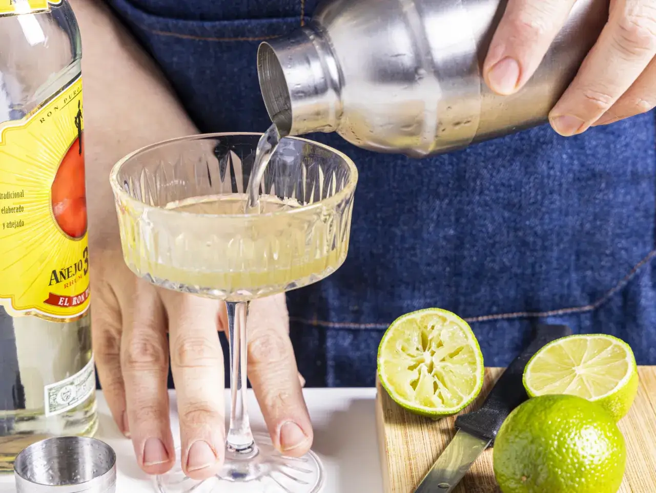 Zubereitung von Daiquiri-Rezepten: Rum wird aus einem Shaker in ein Glas gegossen. Limetten und ein Messer liegen bereit.