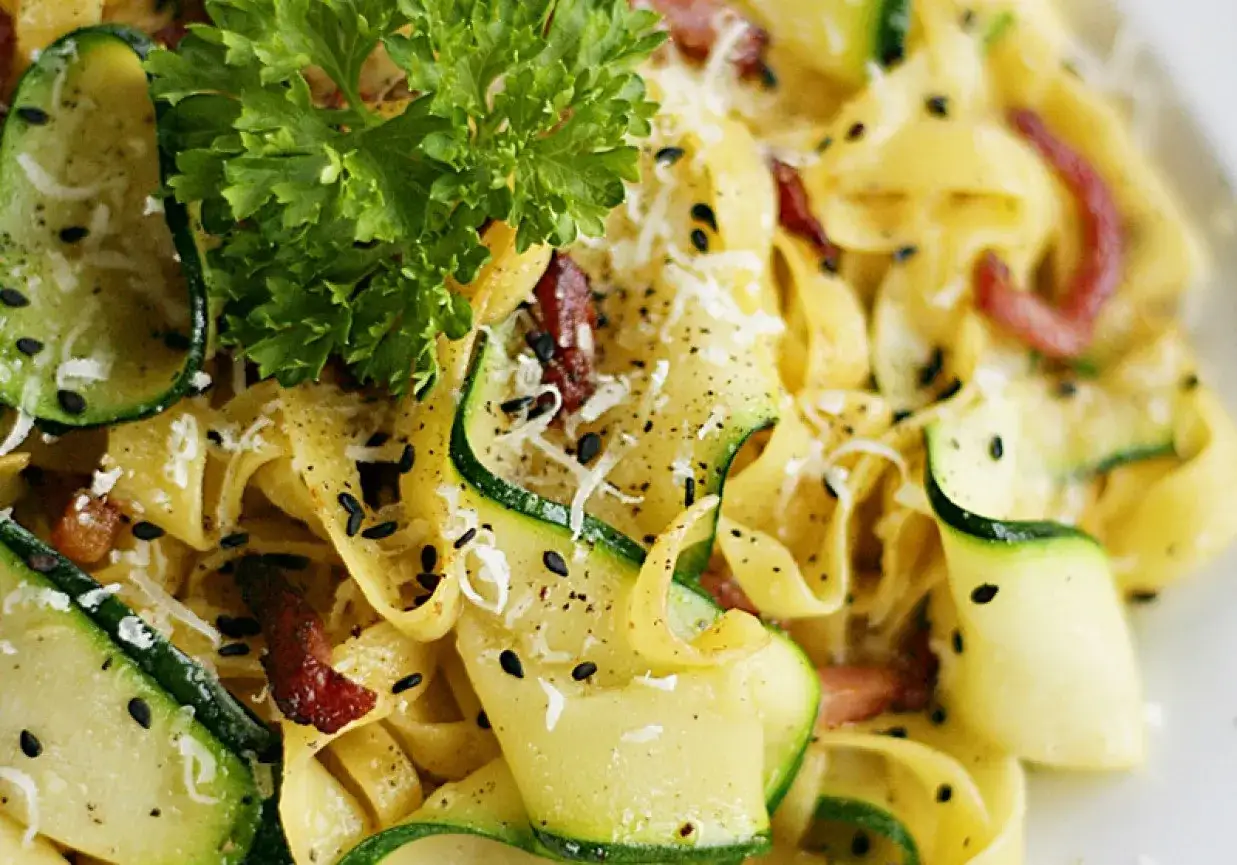 Pyszny makaron z cukinią i boczkiem, posypany parmezanem i czarnym sezamem, ozdobiony gałązką natki pietruszki.