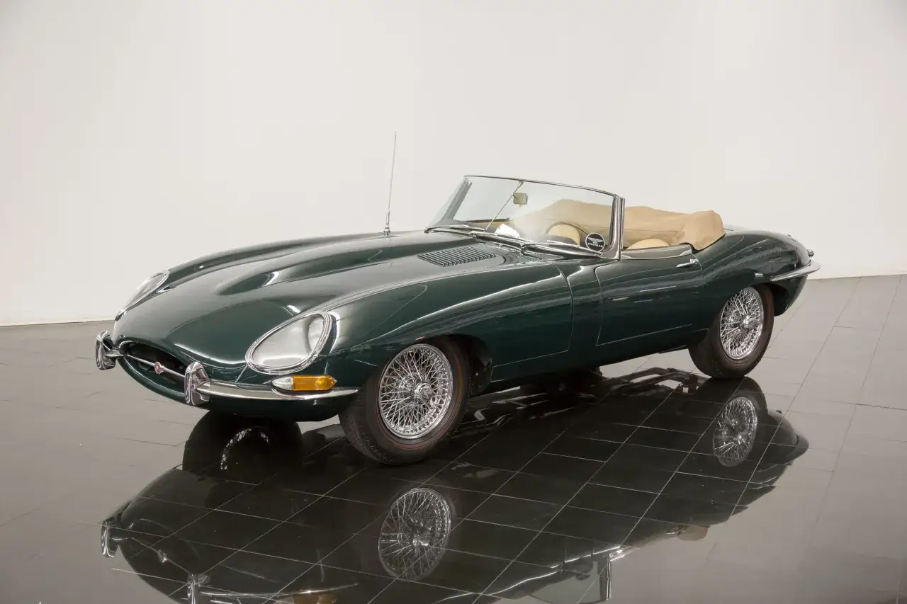 Klasyczny zielony Jaguar E-Type z beżowym dachem i wnętrzem, lśniący na czarnej, lustrzanej podłodze.