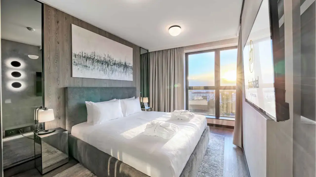 Przytulny pok&oacute;j w Focus Hotel Premium Warszawa. Duże ł&oacute;żko z białymi szlafrokami, nowoczesny wystr&oacute;j i widok na zach&oacute;d słońca.