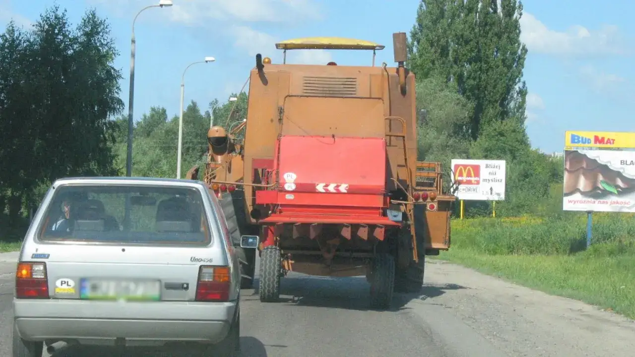 Pomarańczowy pojazd wolnobieżny, prawdopodobnie kombajn, jedzie drogą za srebrnym Fiatem.