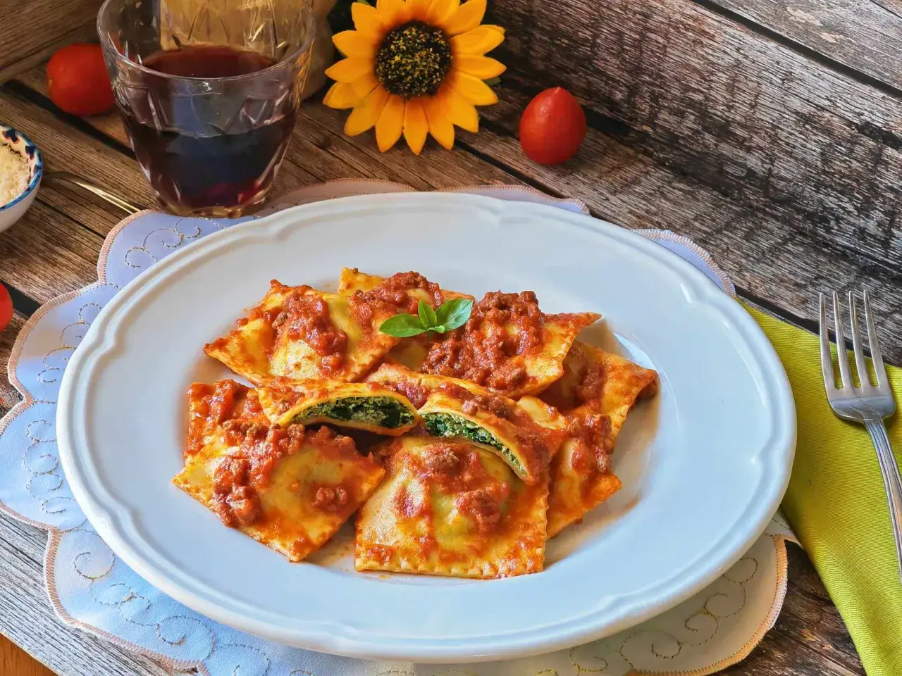 Piatto di ravioli ricotta e spinaci conditi con sugo rosso e ragù, guarniti con basilico. Accanto, un bicchiere di vino rosso e un girasole.