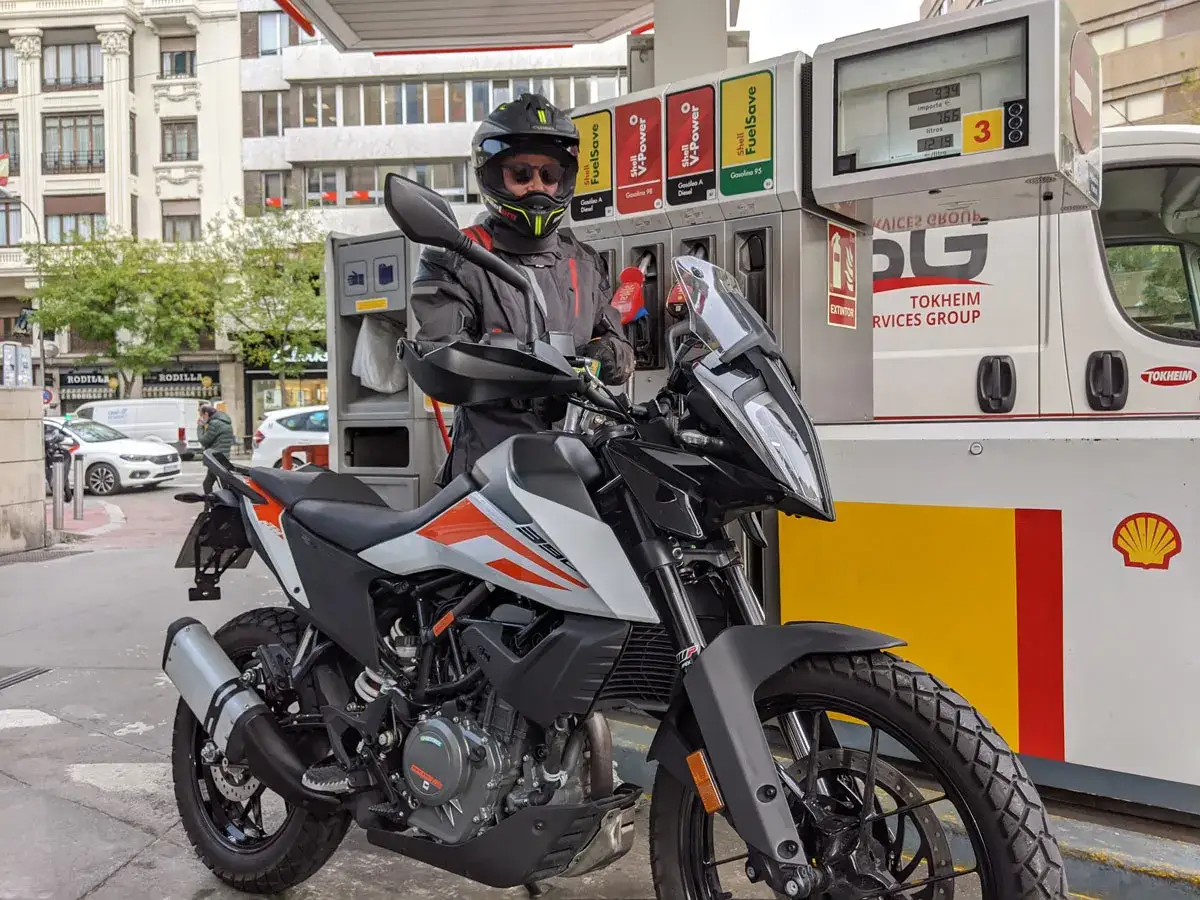 Motociclista en gasolinera, eligiendo entre gasolina 95 y 98 para su moto. El surtidor muestra precios y litros.