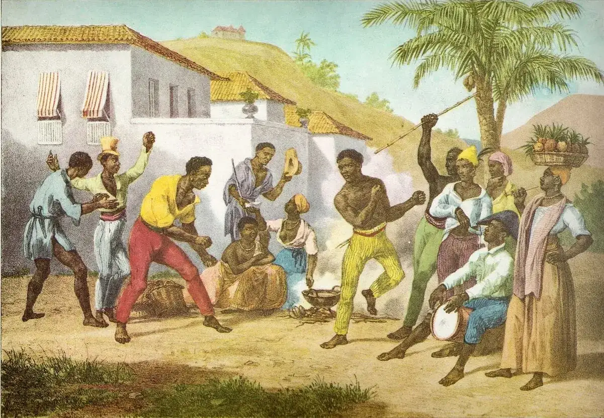 Grupa ludzi tańczy i śpiewa, tworząc rytm do capoeira. Jedna osoba gra na bębnie, inni klaszczą.
