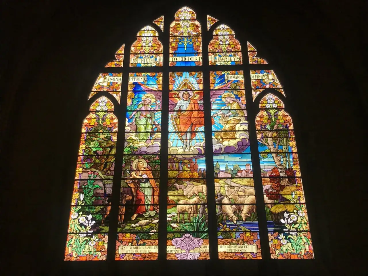 Vitrail de Jeanne d'Arc : une scène céleste avec des anges et une figure centrale en armure, entourée de moutons et d'un paysage bucolique.