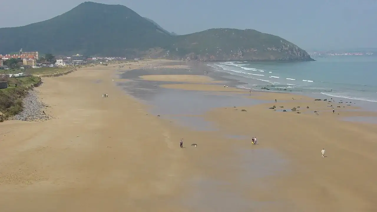 Amplia vista de la playa de Berria con gente paseando y monta&ntilde;as verdes al fondo.