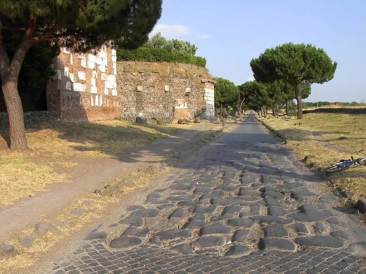 Starożytna Via Appia, brukowana droga z kamienia, otoczona drzewami i ruinami.