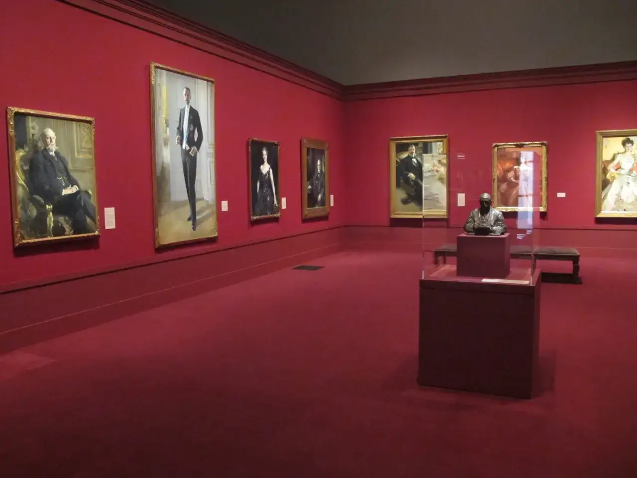 En utställningssal med röda väggar och matta. Porträtt av män och kvinnor hänger på väggarna, kanske inspirerade av anders zorn film. En byst står på ett podium.