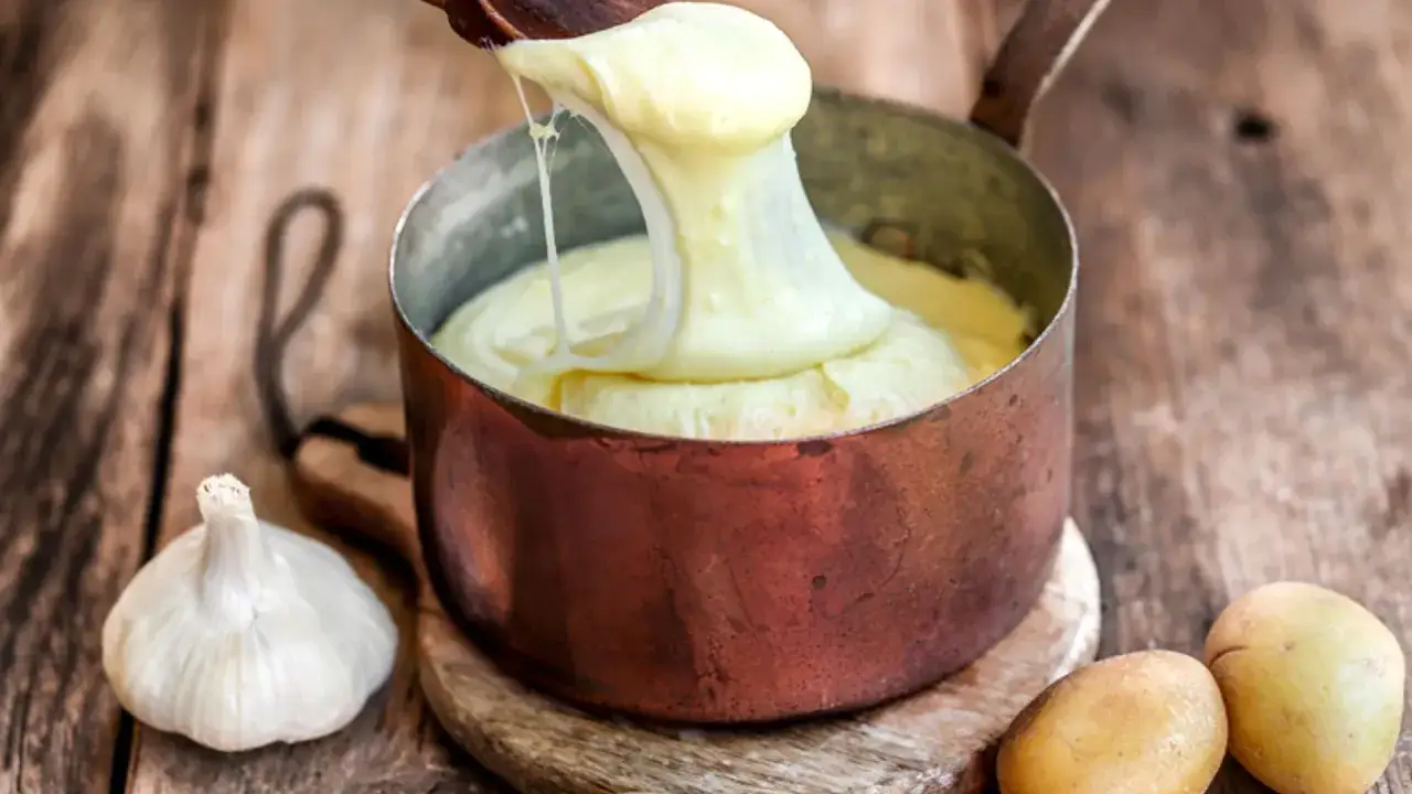 Aligot receta: pur&eacute; de patatas cremoso y el&aacute;stico servido en una olla de cobre, con ajos y patatas crudas al lado.
