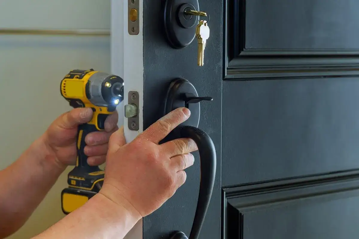 Mains tenant une perceuse pour changer la serrure d'une porte noire. Une clé est insérée dans la nouvelle serrure.