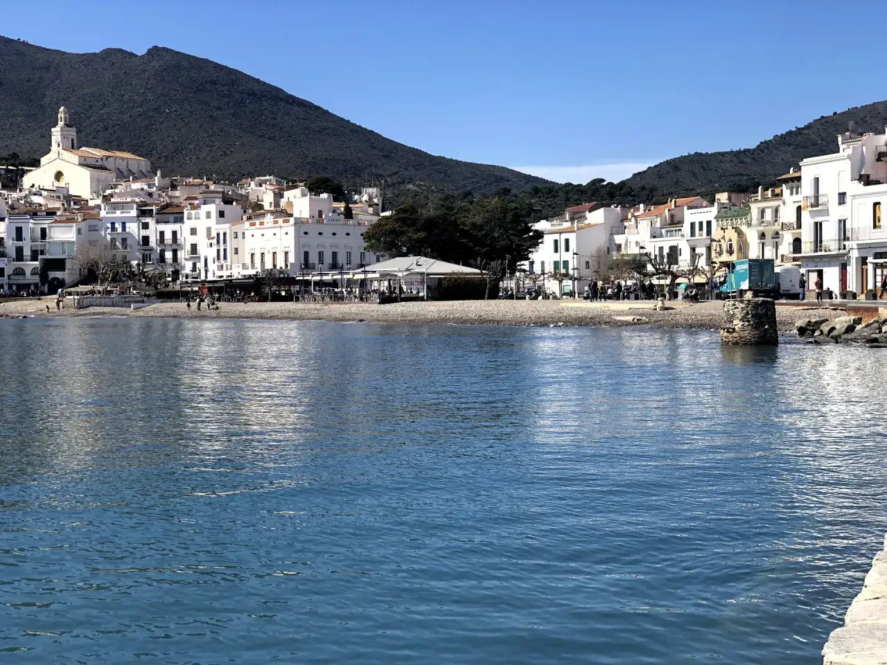 Vista del mar azul frente a la platja gran Cadaqu&eacute;s, con casas blancas y una iglesia en la ladera de una monta&ntilde;a verde.