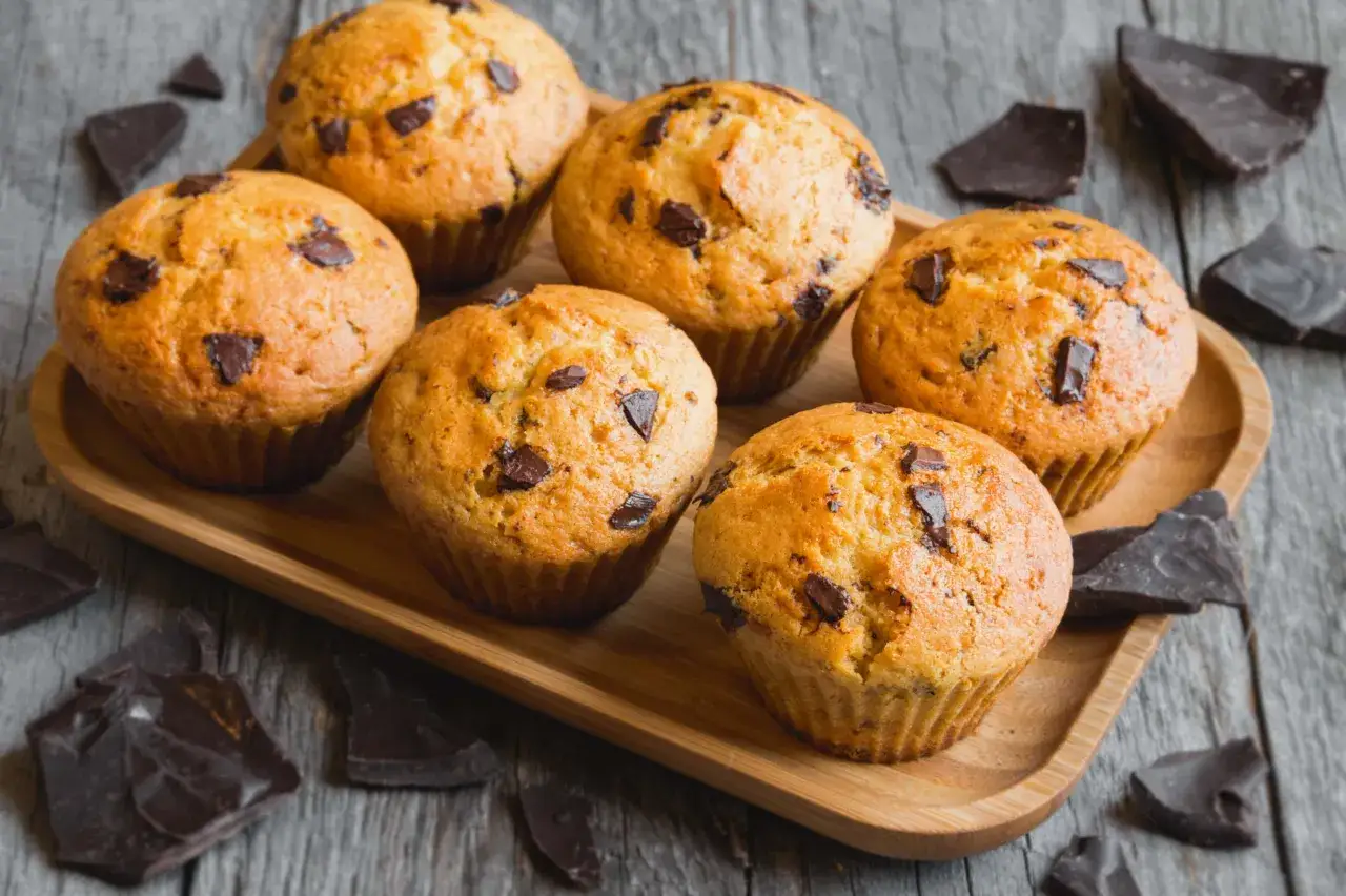 Sei muffin allo yogurt con gocce di cioccolato su un vassoio di legno, circondati da pezzi di cioccolato fondente.