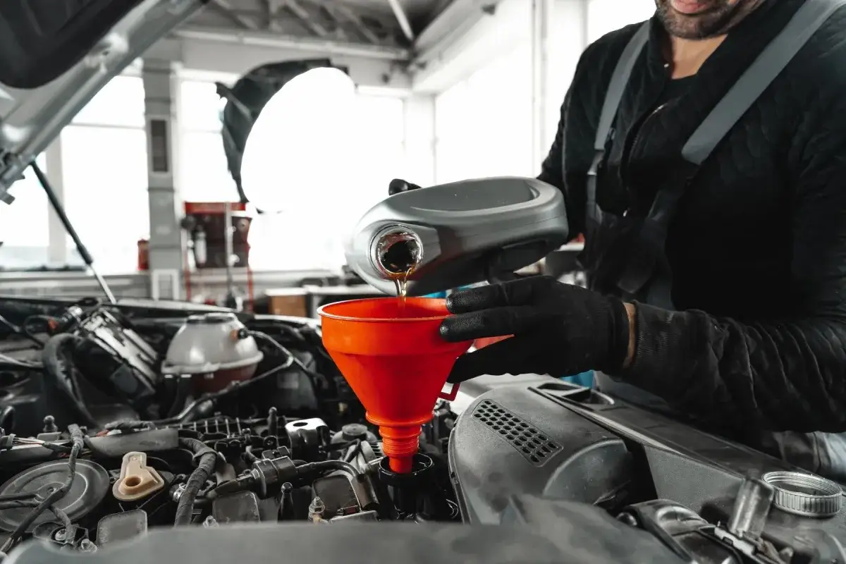 Mechanik w czarnej bluzie i rękawiczkach wlewa olej do silnika przez czerwony lejek. To kluczowy etap płukania silnika przed wymianą oleju.