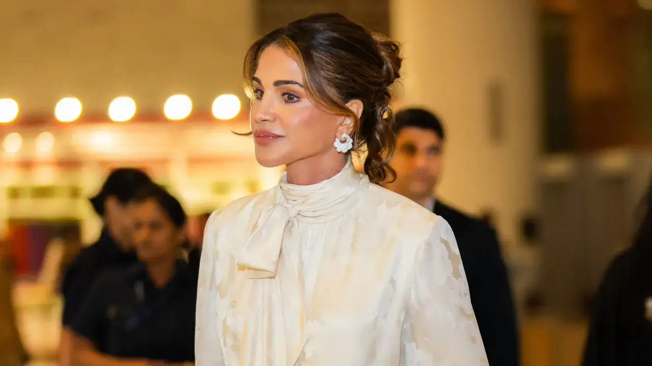 A woman in a cream-colored blouse with a bow, embodying queen rania style, wears floral earrings.