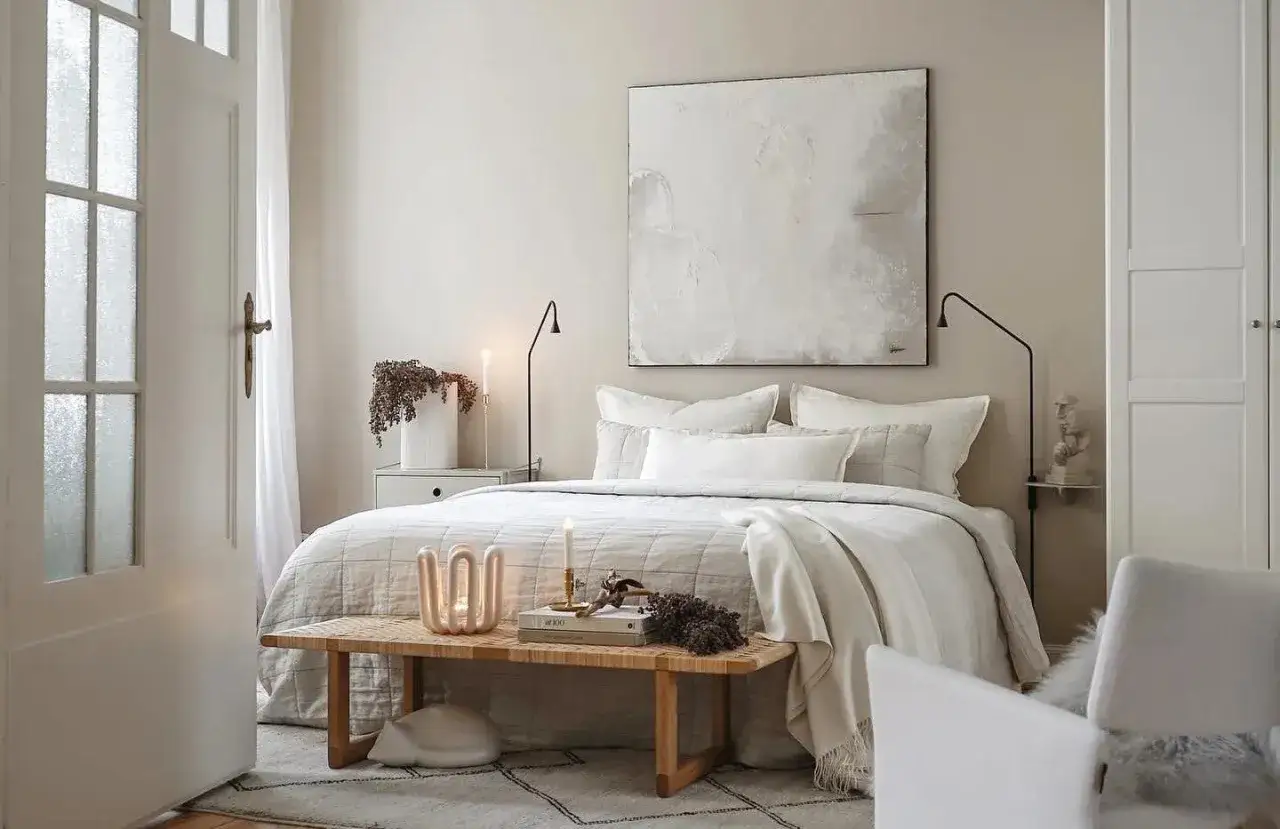 A serene bedroom with neutral interior design. A large abstract painting hangs above a bed dressed in soft, light linens. A wooden bench with decorative items sits at the foot of the bed.