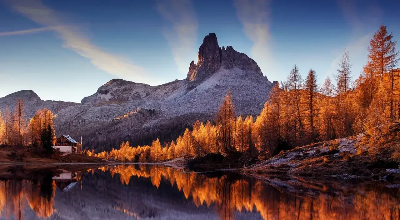 Riflessi dorati sull'acqua di un lago alpino, con un rifugio e montagne innevate. Perfetto per le camminate a Cortina d'Ampezzo.