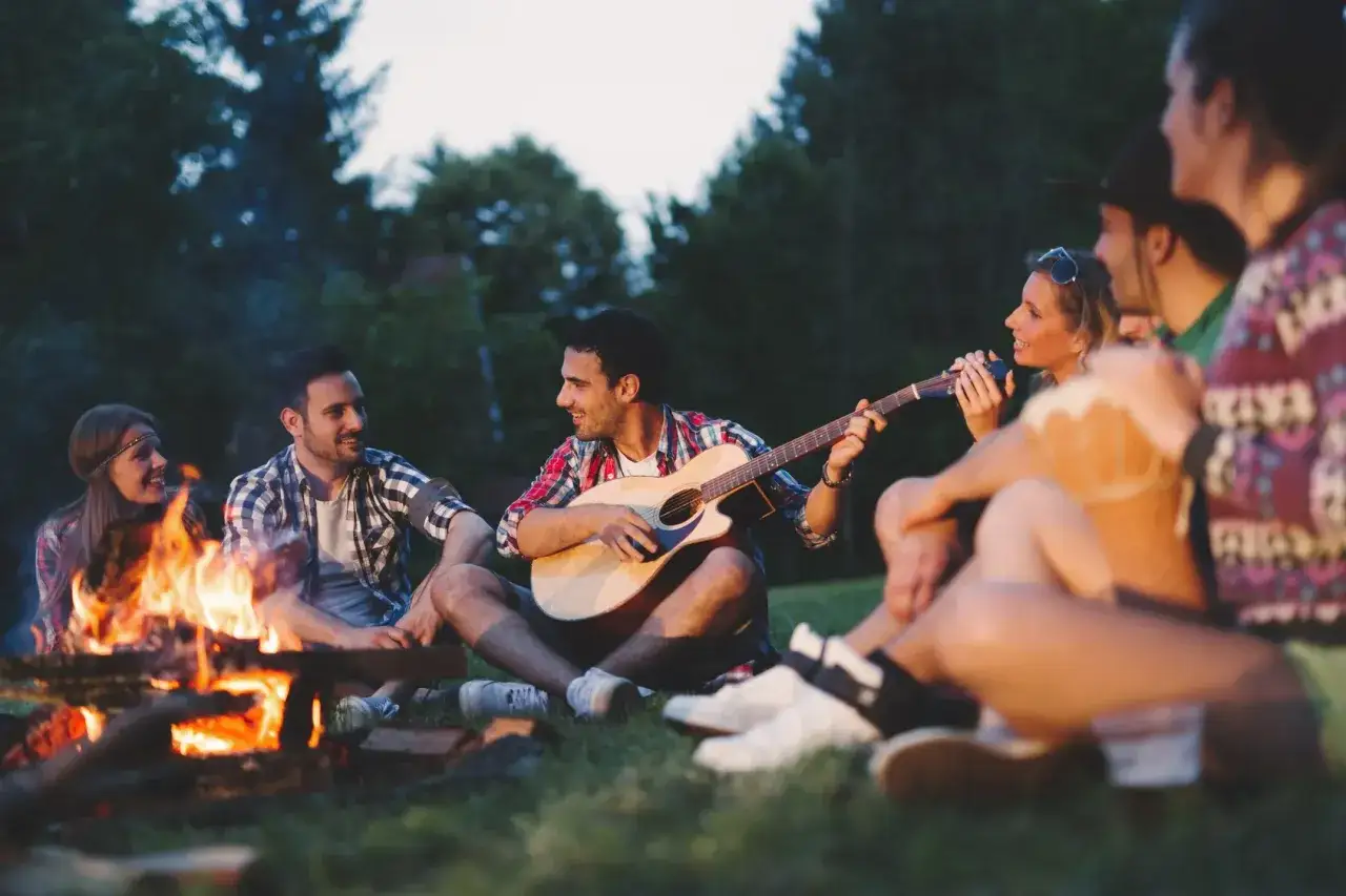 Przy ognisku rozbrzmiewają znane piosenki. Grupa przyjaciół siedzi na trawie, jeden gra na gitarze, inni śpiewają i śmieją się.