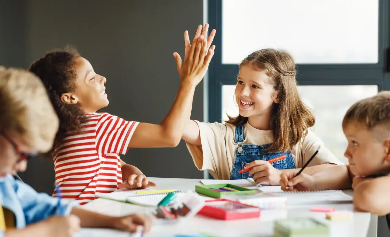 Zwei Kinder geben sich die Hand &uuml;ber einem Tisch voller Lernmaterialien. Ein Zeichen f&uuml;r guten Unterricht und Erfolg.