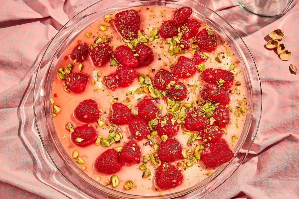 En krämig panna cotta med hallon och pistagenötter i en glasform, serverad på en rosa duk.