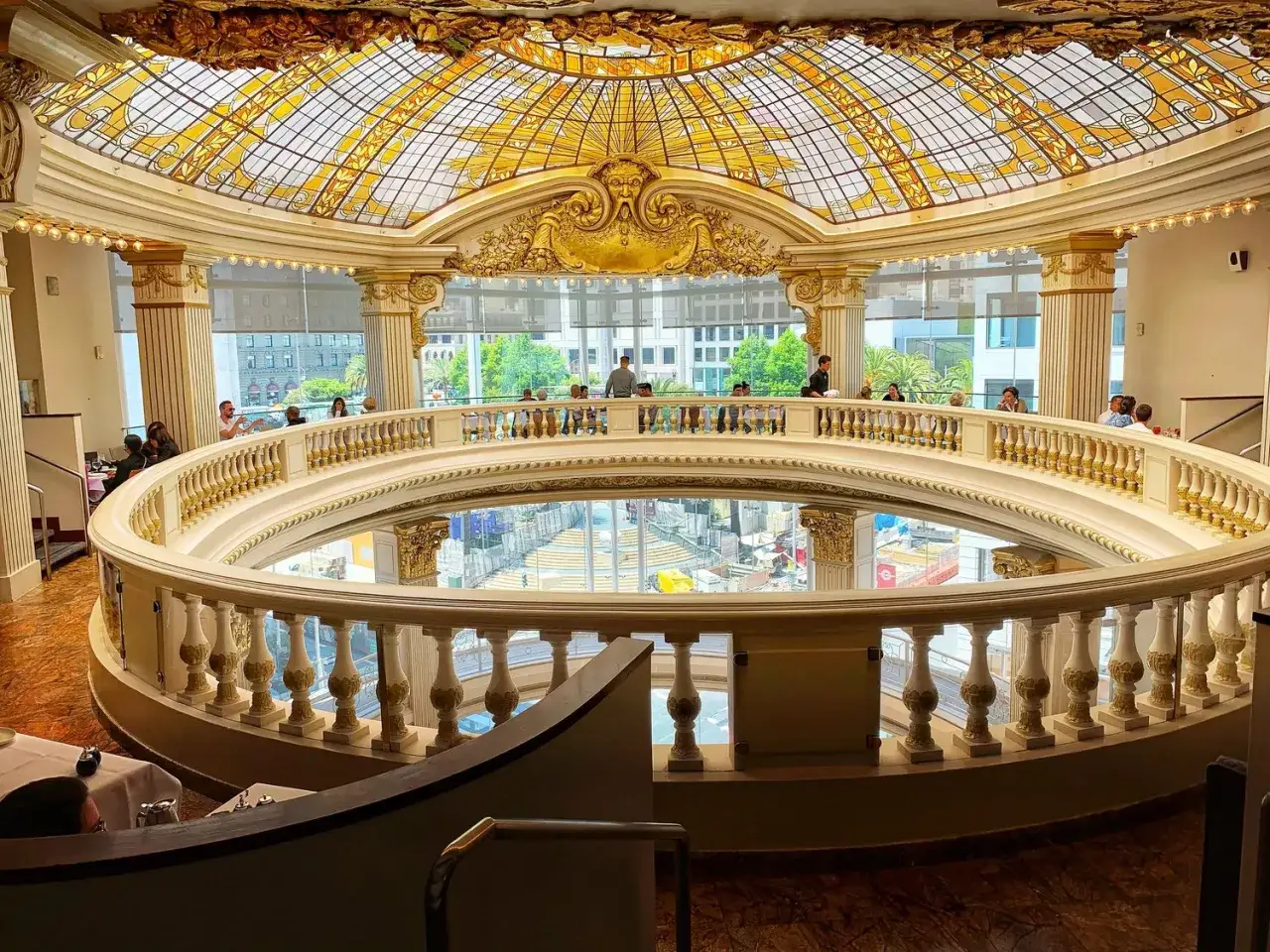 Elegancka rotunda z witrażowym sufitem i złotymi zdobieniami. Ludzie siedzą przy balustradzie, podziwiając widok.