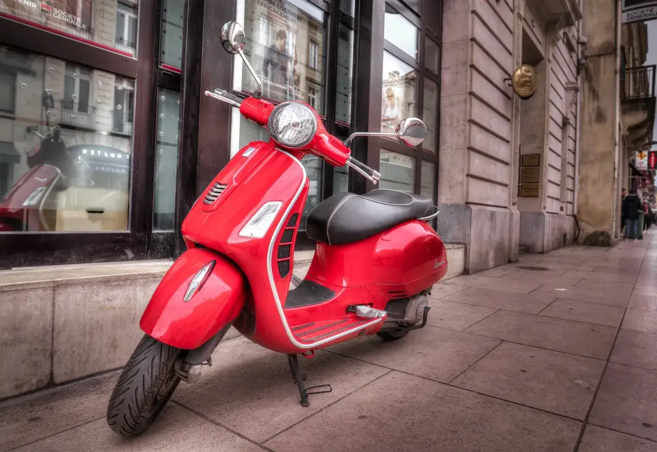 Czerwona Vespa na chodniku. Czy na motorynkę trzeba mieć prawo jazdy? To pytanie zadaje sobie wielu, widząc taki skuter.