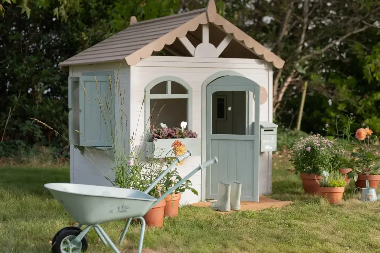 Ein bezauberndes Spielhaus, das zum **Spielhaus selber bauen** inspiriert. Mit Schubkarre, Gummistiefeln und Blumenk&auml;sten wirkt es wie ein kleines Paradies im Garten.