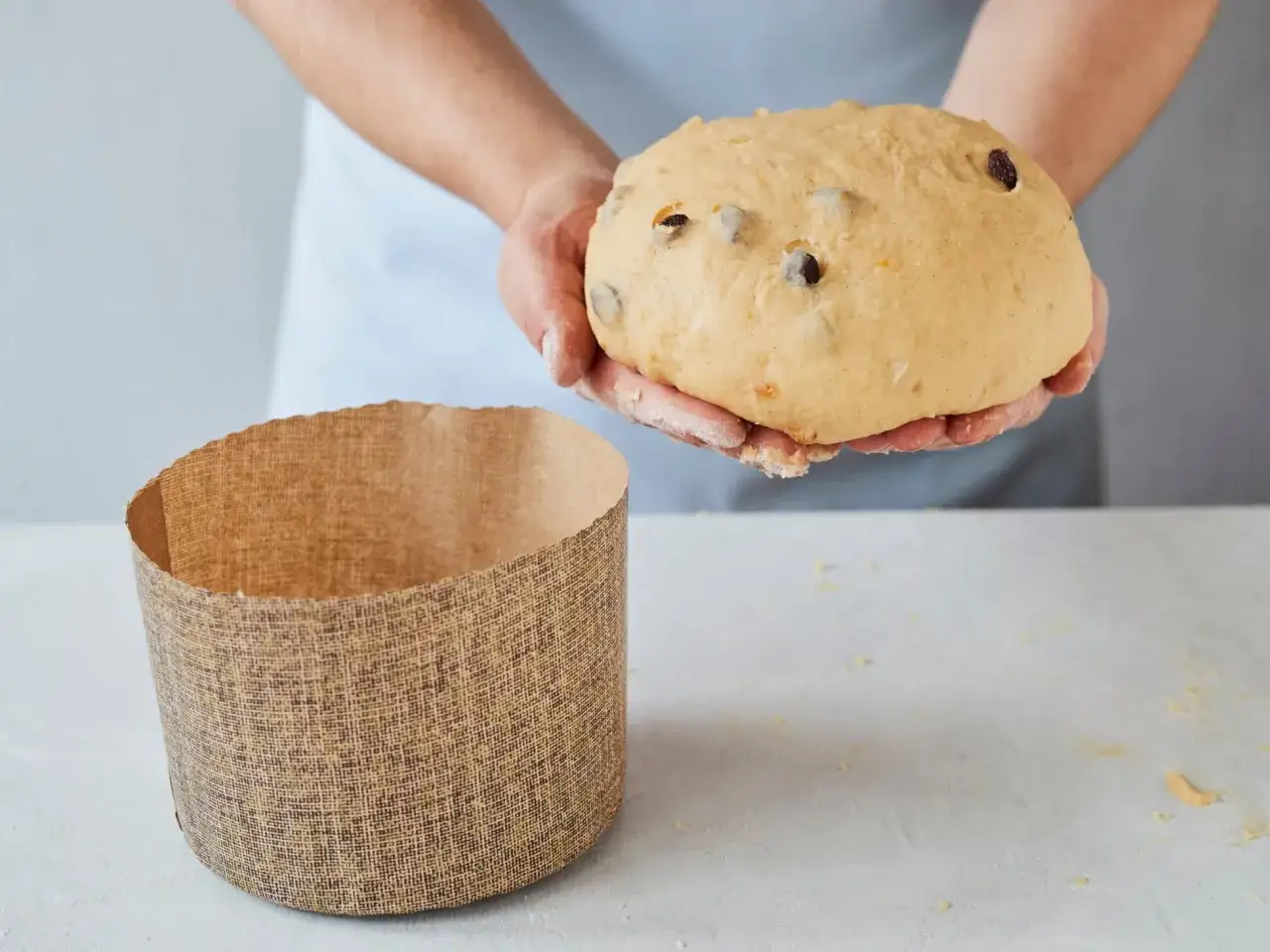 Hände halten einen Teig für ein Panettone Rezept, daneben steht eine leere Backform.