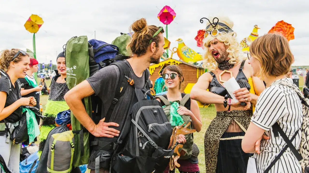 Festival-goers with backpacks chat near colorful decorations. One person in drag checks a festival check list.