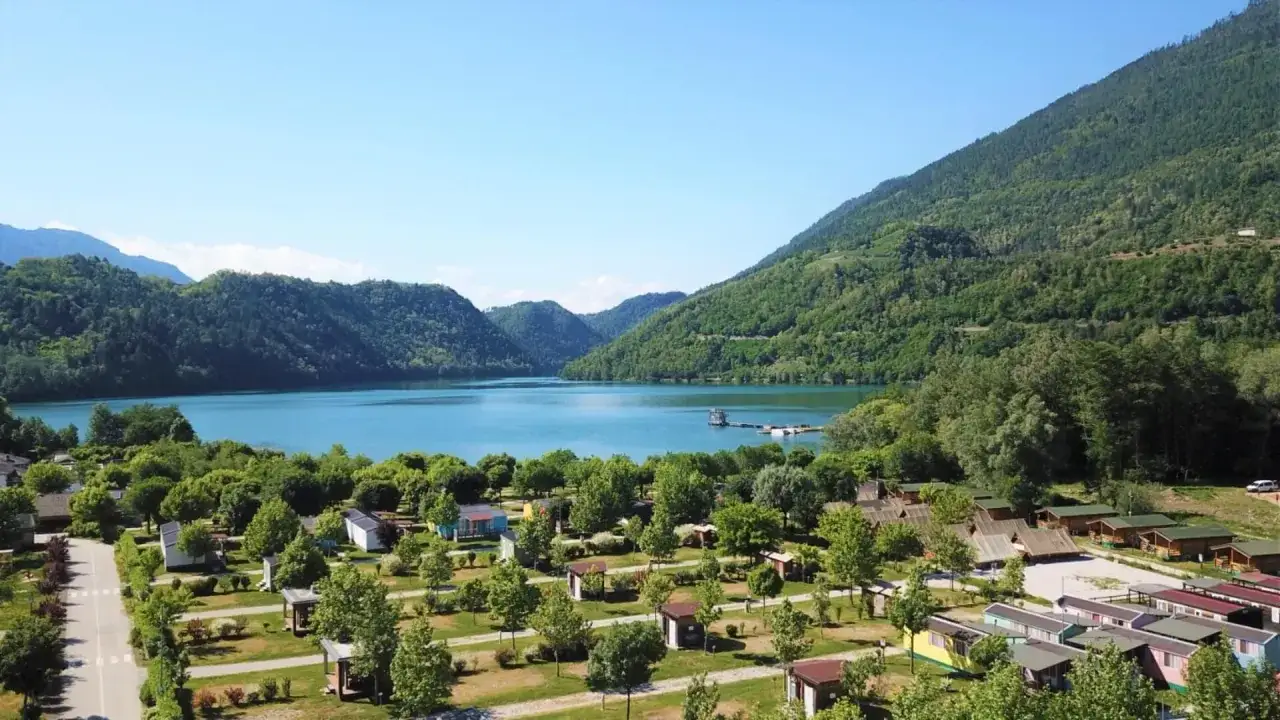 Un incantevole **lago Levico camping village** immerso nel verde, con bungalow colorati e il lago azzurro che si estende tra le montagne.