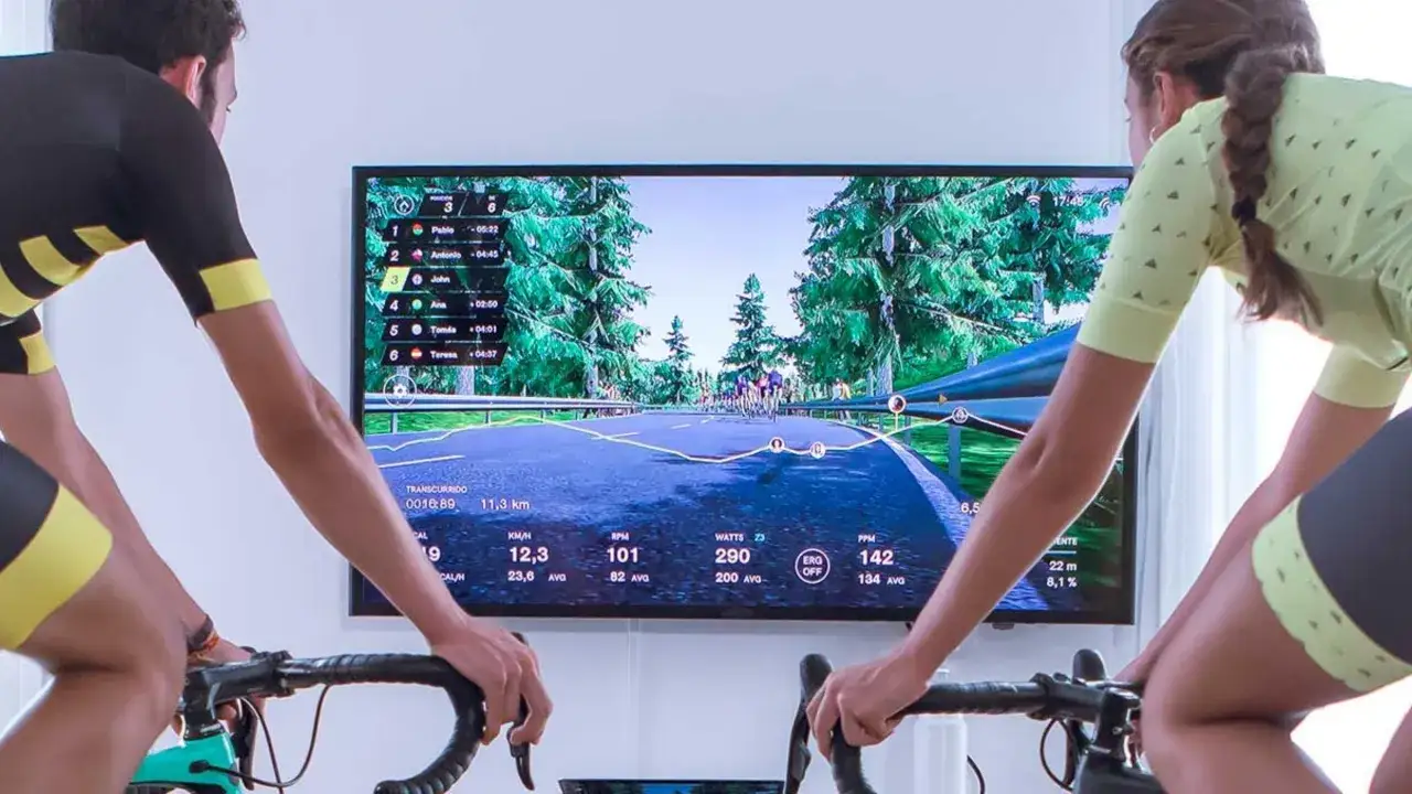 Two cyclists race virtually on a scenic road using indoor cycling apps, displaying real-time stats on a large screen.
