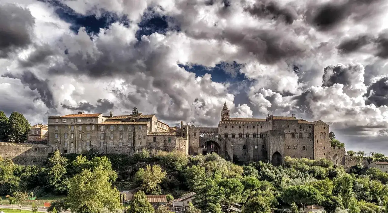 Antico borgo medievale in **Tuscia**, dove si trova questo imponente complesso di edifici storici su una collina verdeggiante, sotto un cielo drammatico.