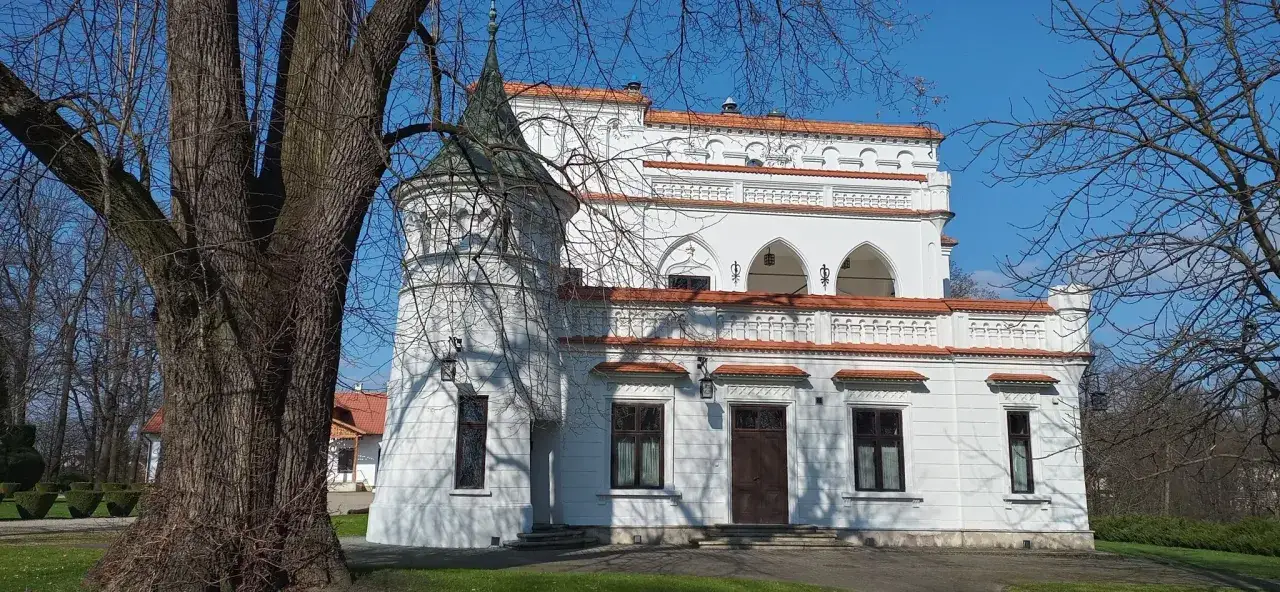 Biały pałac z wieżyczką i czerwonym dachem, otoczony drzewami. W oddali widać inne budynki. To piękny **Radomyśl Wielki**.