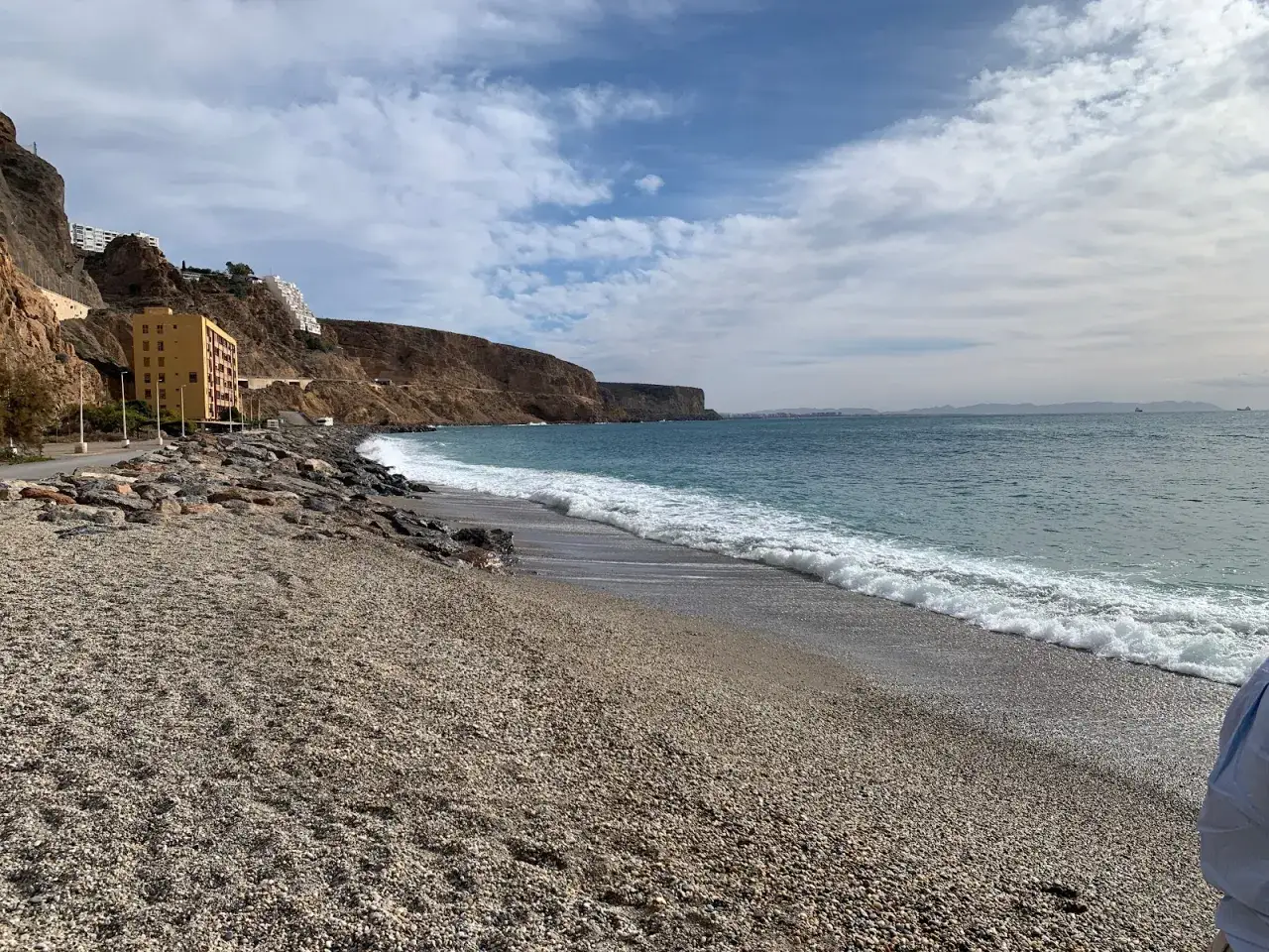 La playa de guijarros de el palmer almeria, con olas rompiendo suavemente y edificios en la ladera rocosa.