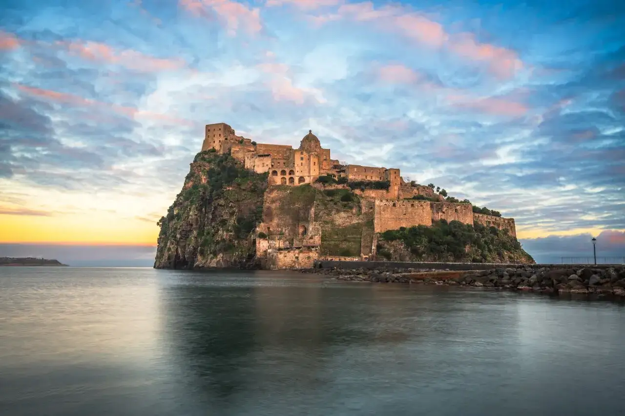 Castello Aragonese su uno scoglio roccioso, con il mare calmo e un cielo colorato al tramonto. Ispirazione per frasi su Ischia.