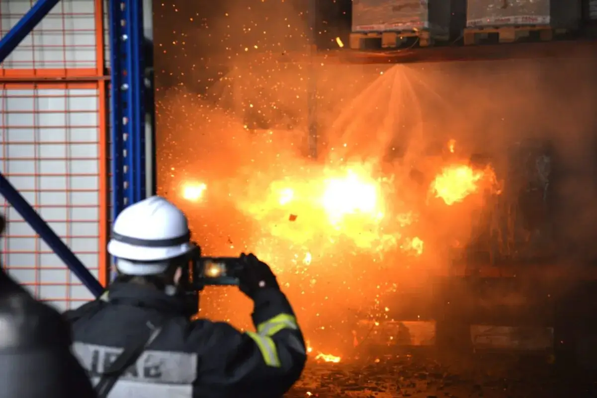 Feuerwehrleute dokumentieren eine Explosion, die die **Brandlast**-Definition veranschaulicht. Funken und Flammen lodern in einem Lager.