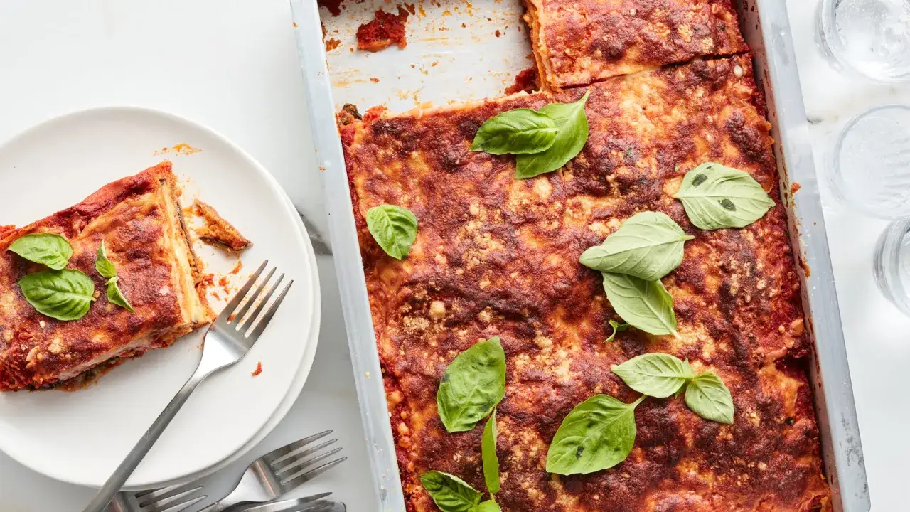 Un plat d'aubergine parmigiana tout juste sorti du four, garni de feuilles de basilic frais. Une part est servie sur une assiette blanche avec des couverts.
