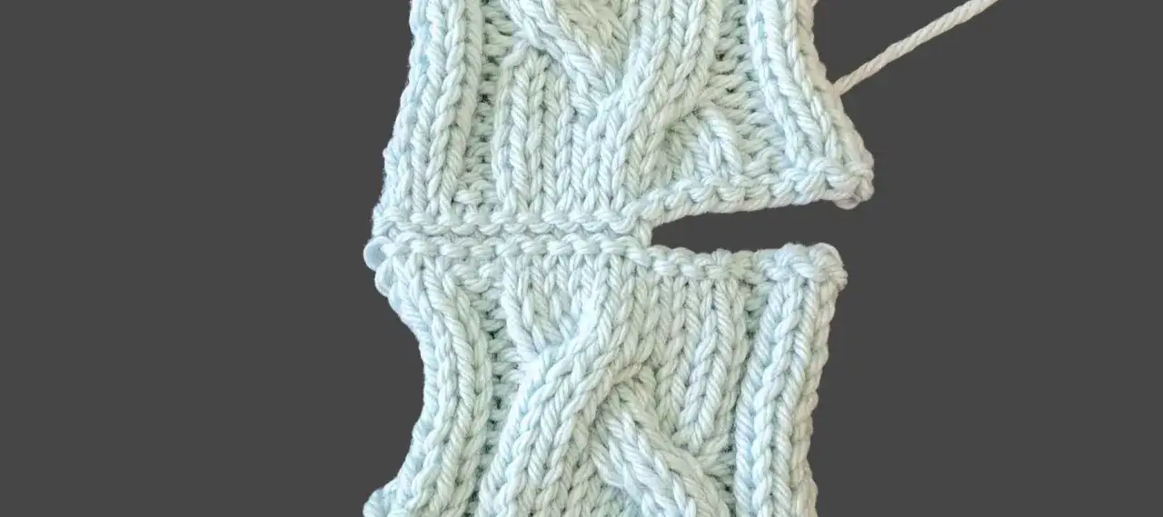 Close-up of light blue knitted fabric, showing the process of sewing shoulder seams. The cable knit pattern is visible.