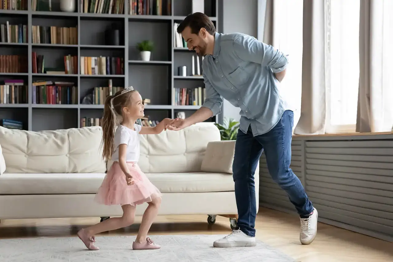 Padre y hija bailan, ella con tiara y falda rosa, él sonríe. Parecen decirse palabras mágicas en su juego.