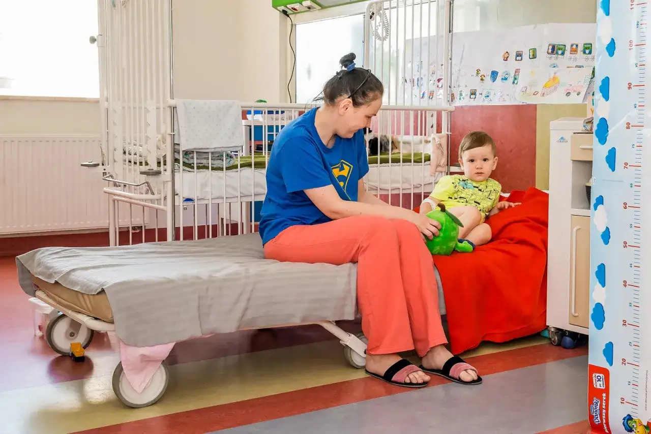 Mama opiekuje się dzieckiem w szpitalu. Maluch siedzi na łóżku, przytulając zieloną zabawkę.