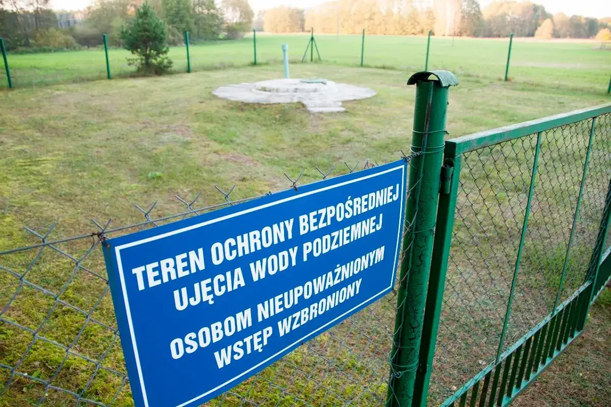 Niebieska tablica informuje o **strefach ochronnych** ujęcia wody podziemnej. Teren ogrodzony, wstęp wzbroniony.