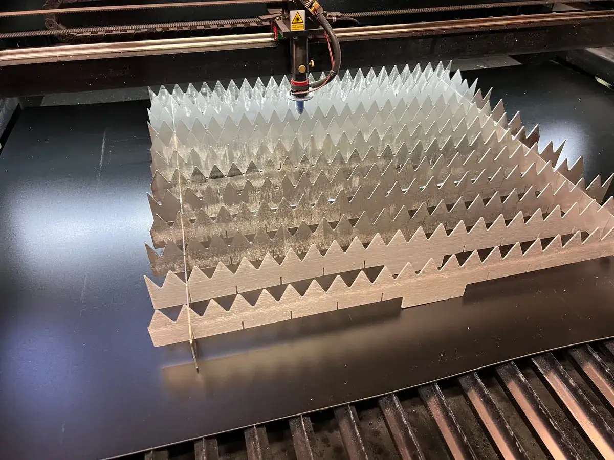 A laser cutter is in action, demonstrating how to laser cut acrylic at home. The machine precisely cuts a metallic sheet into a zig-zag pattern.