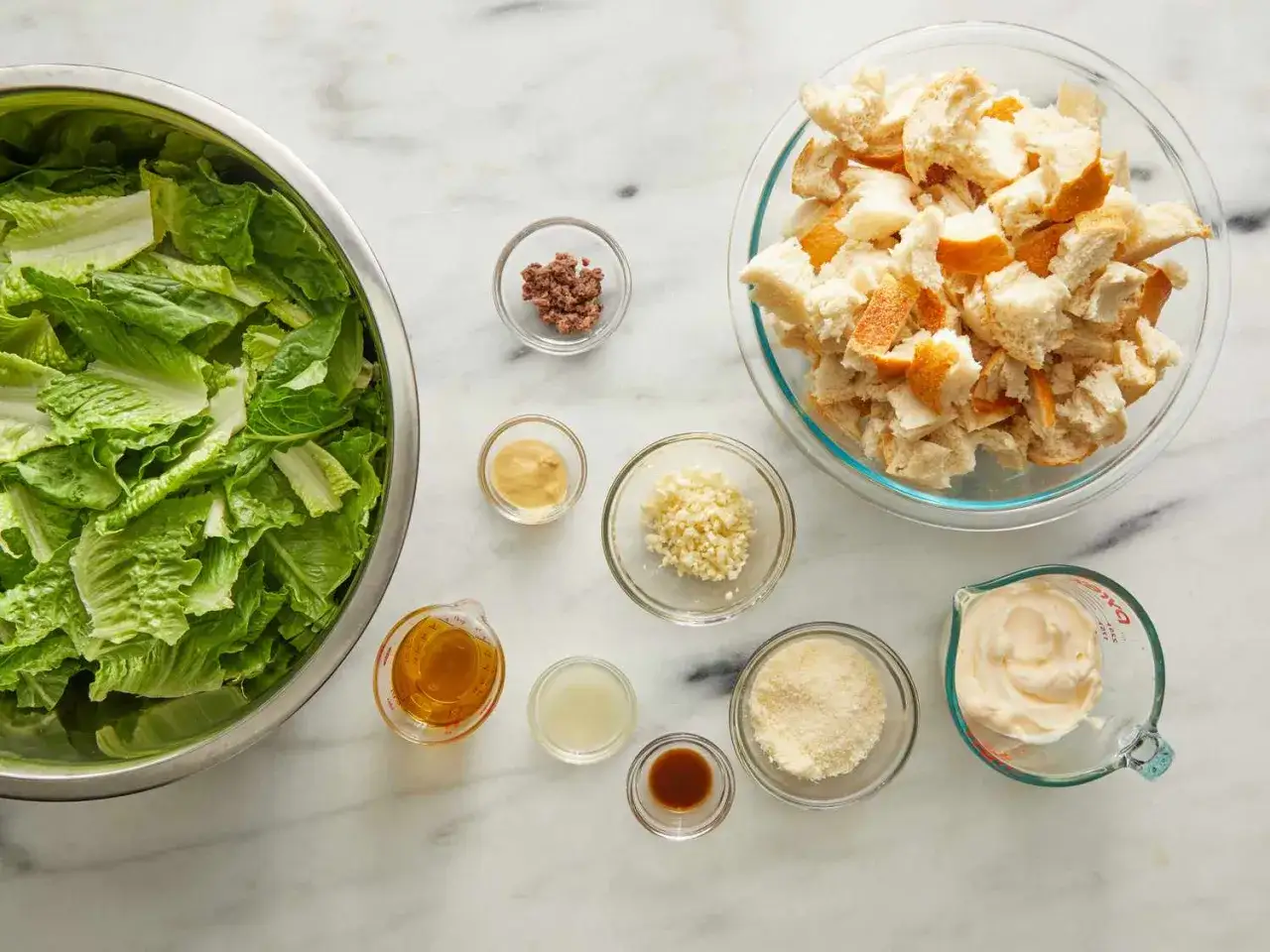 Alla ingredienser för ett klassiskt caesardressing recept ligger uppradade: romansallad, krutonger, vitlök, parmesan, sardeller, senap, olja, citron och majonnäs.