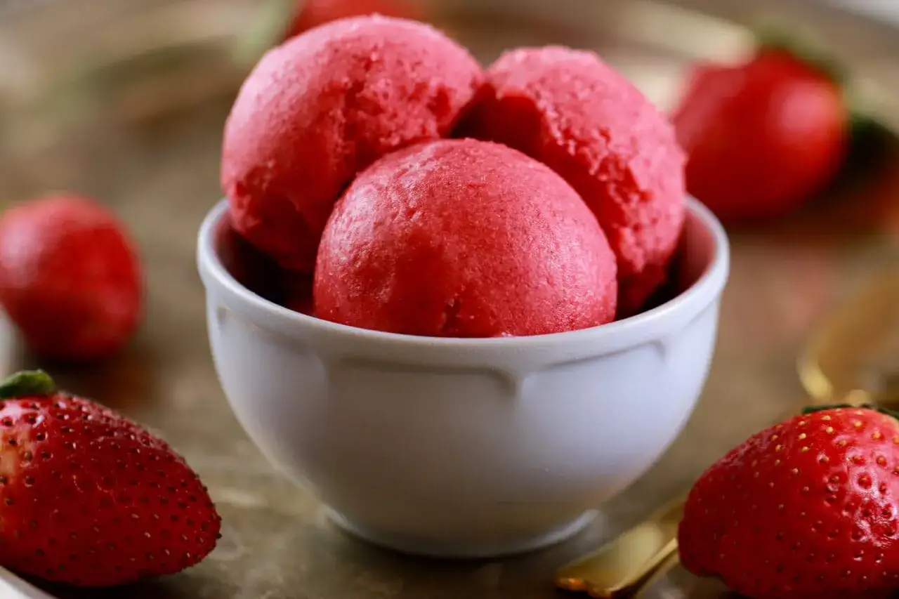 Schüssel mit leuchtend rotem Joghurt-Eis, das zeigt, wie man Joghurt-Eis selber machen ohne Eismaschine kann. Frische Erdbeeren umgeben die Schüssel.
