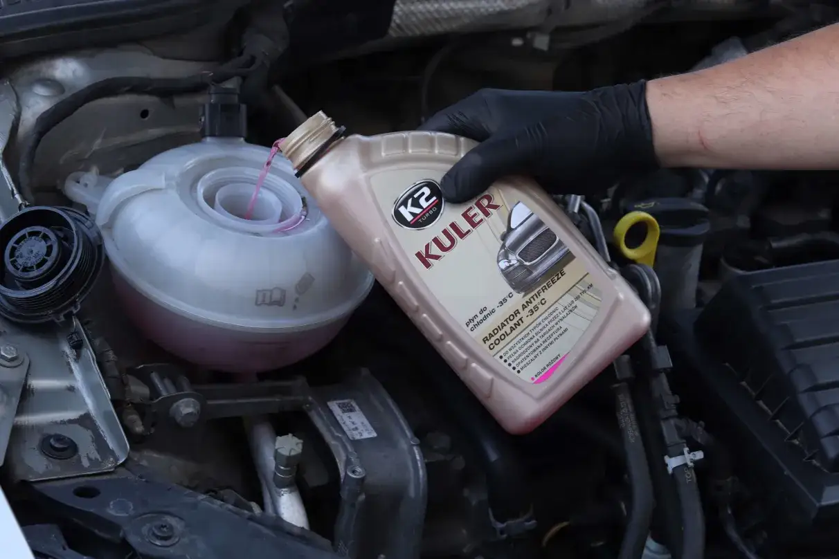 Main d'une personne en gant noir versant du liquide de refroidissement rose dans le réservoir d'une voiture. La question